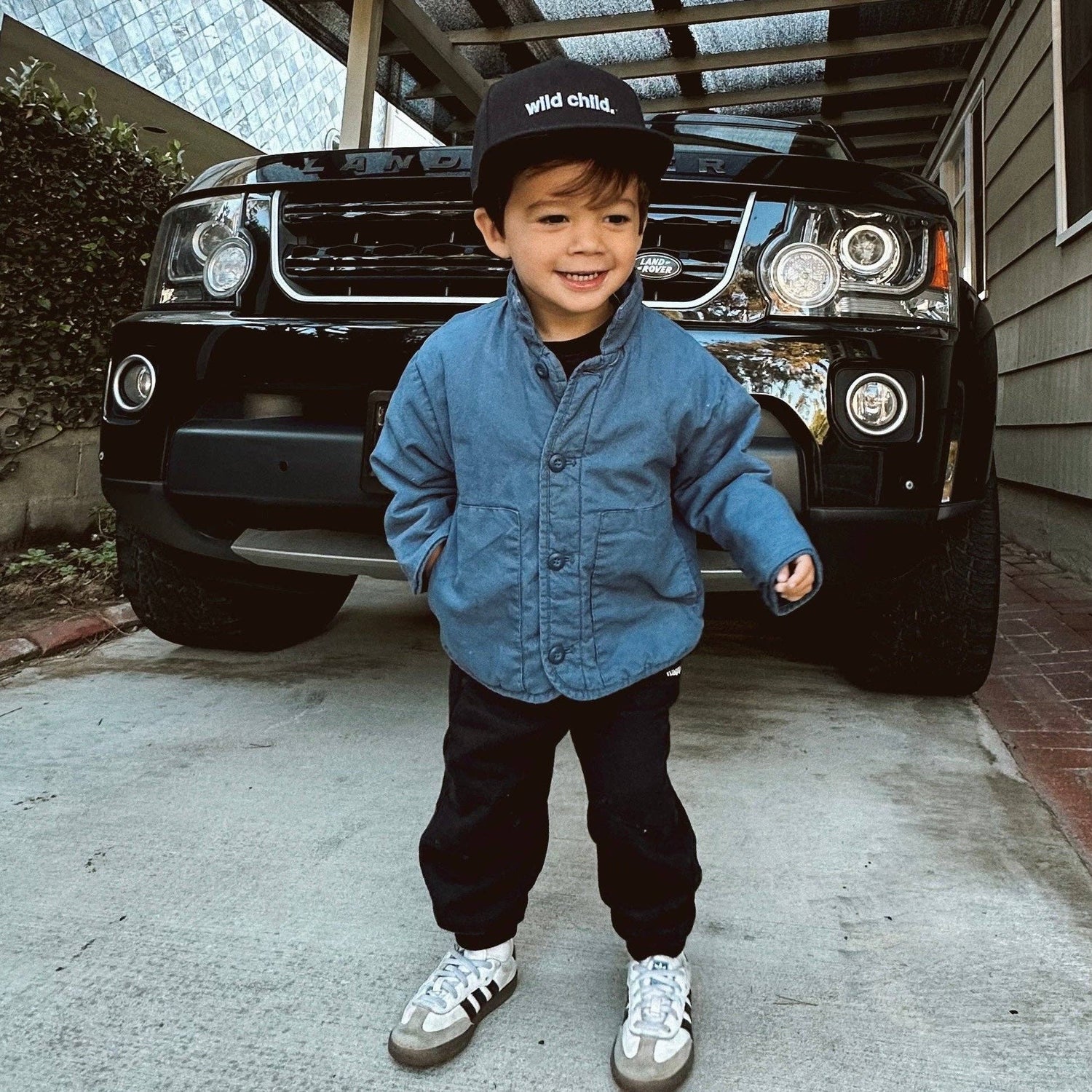 Toddler wearing a toddler snapback hat and stylish jacket, posing beside a black SUV in a driveway.