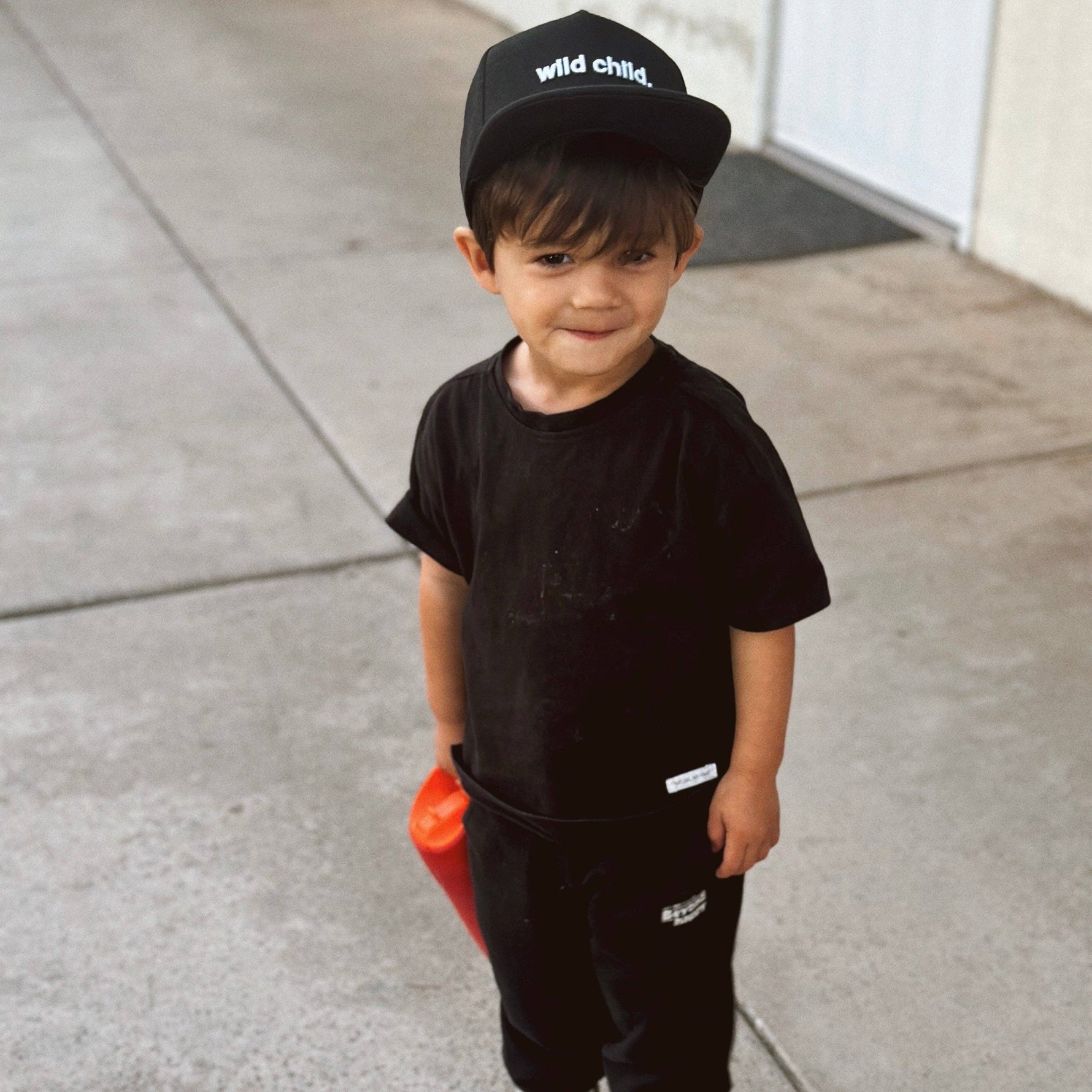 Toddler wearing a black wild child toddler snapback hat and matching outfit while holding a toy.